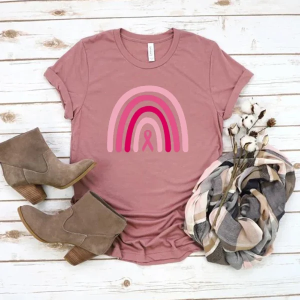 A Rainbow Shirt Pink Ribbon T-shirt with a rainbow and boots on a white wood surface.